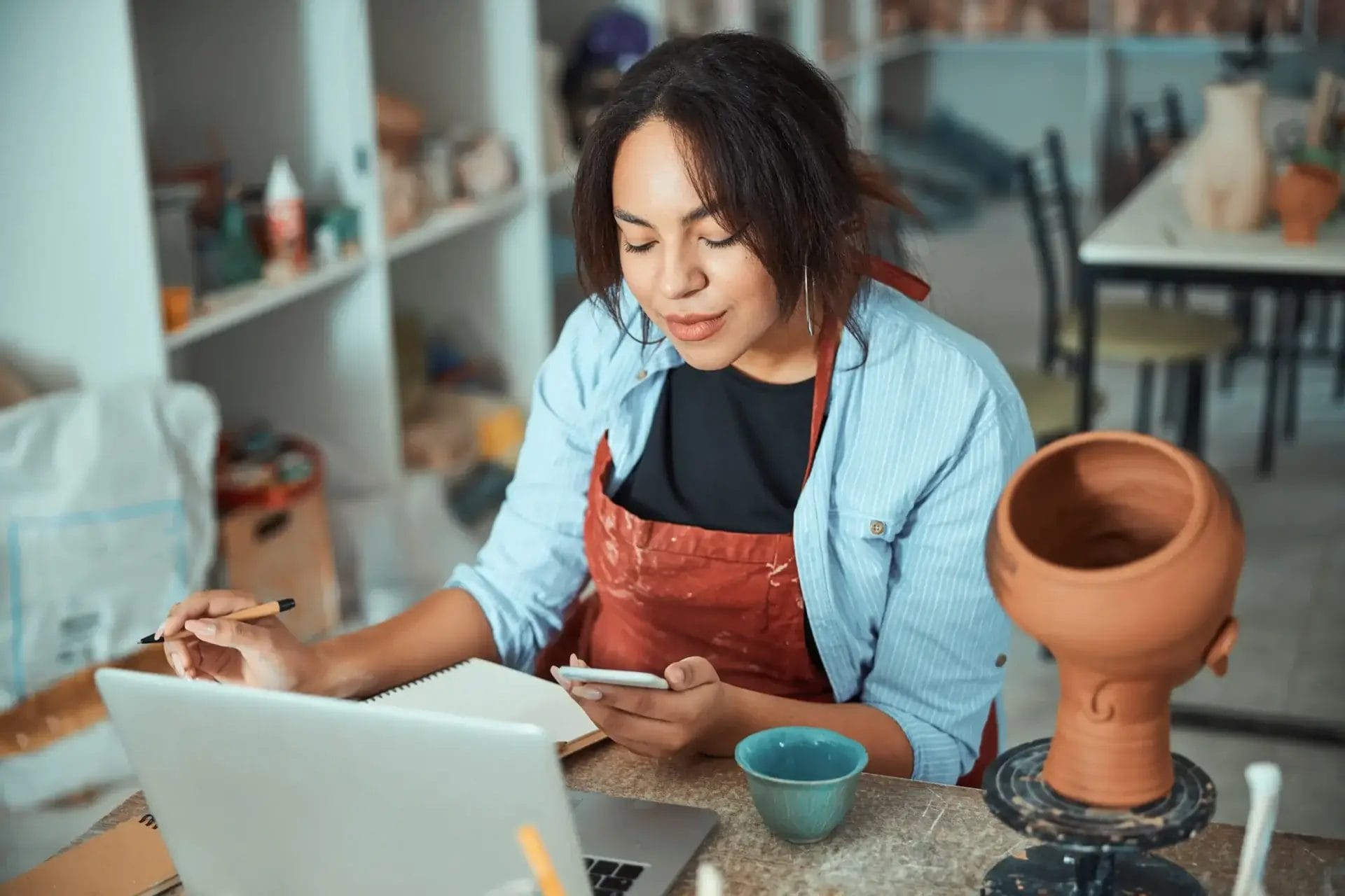 A person in a red apron sits at a table in Charlotte with a laptop, notebook, mug, phone, and handmade pottery piece nearby.