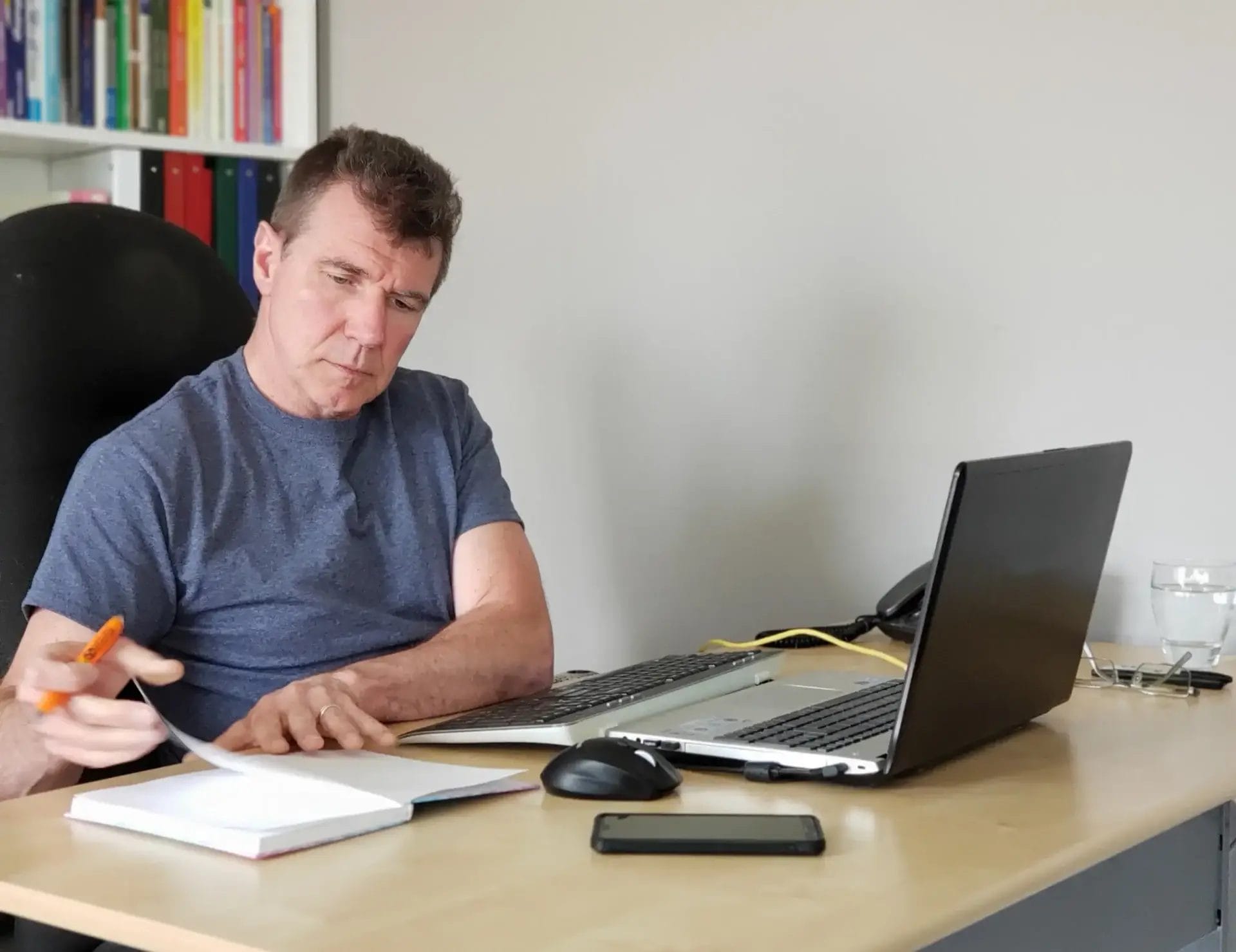 A man writes in a notebook at his desk, laptop open, office supplies nearby, bookshelves behind—working on local Charlotte business ideas.