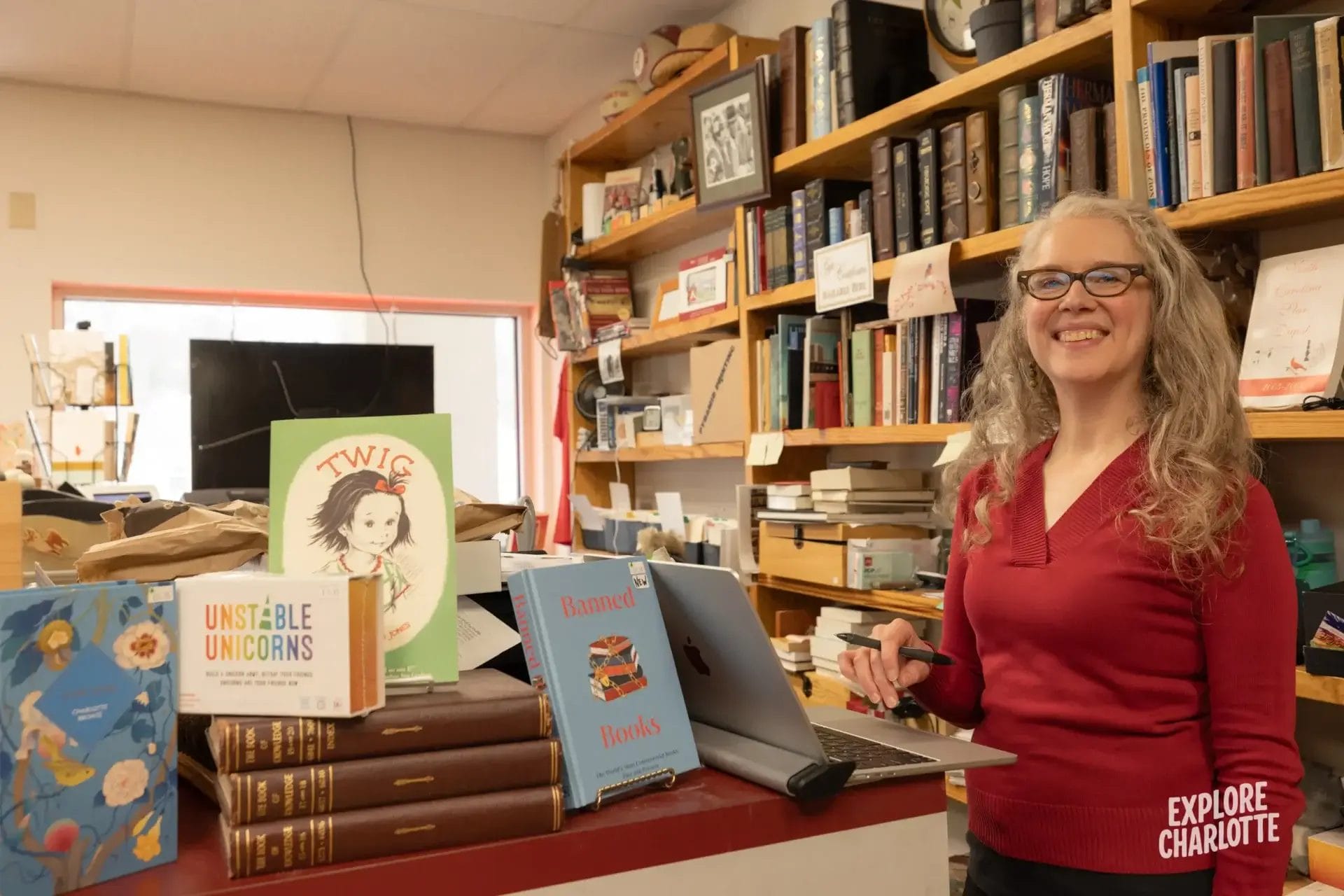 A woman with gray hair and glasses smiles behind a busy bookstore counter in Charlotte, surrounded by books and packages.
