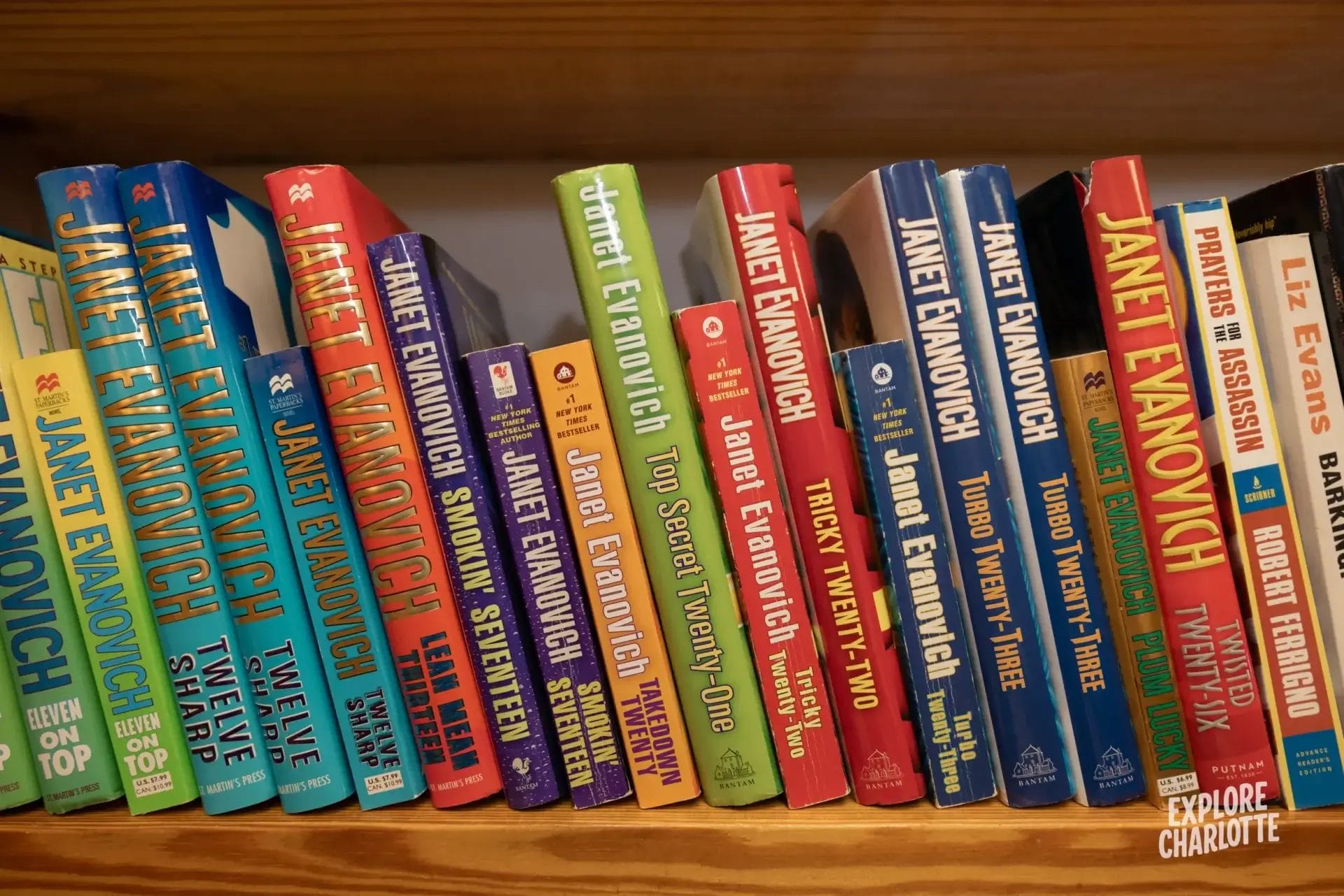 A wooden bookshelf neatly lined with colorful paperbacks, mostly Janet Evanovich novels, like you might see in a Charlotte library.
