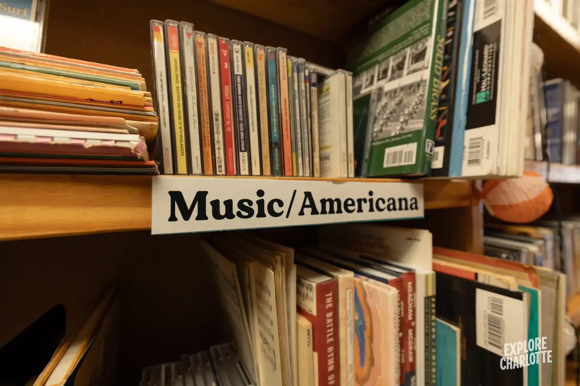 A "Music/Americana" shelf with CDs, books, and records stands in a Charlotte store. Explore Charlotte logo is in the corner.