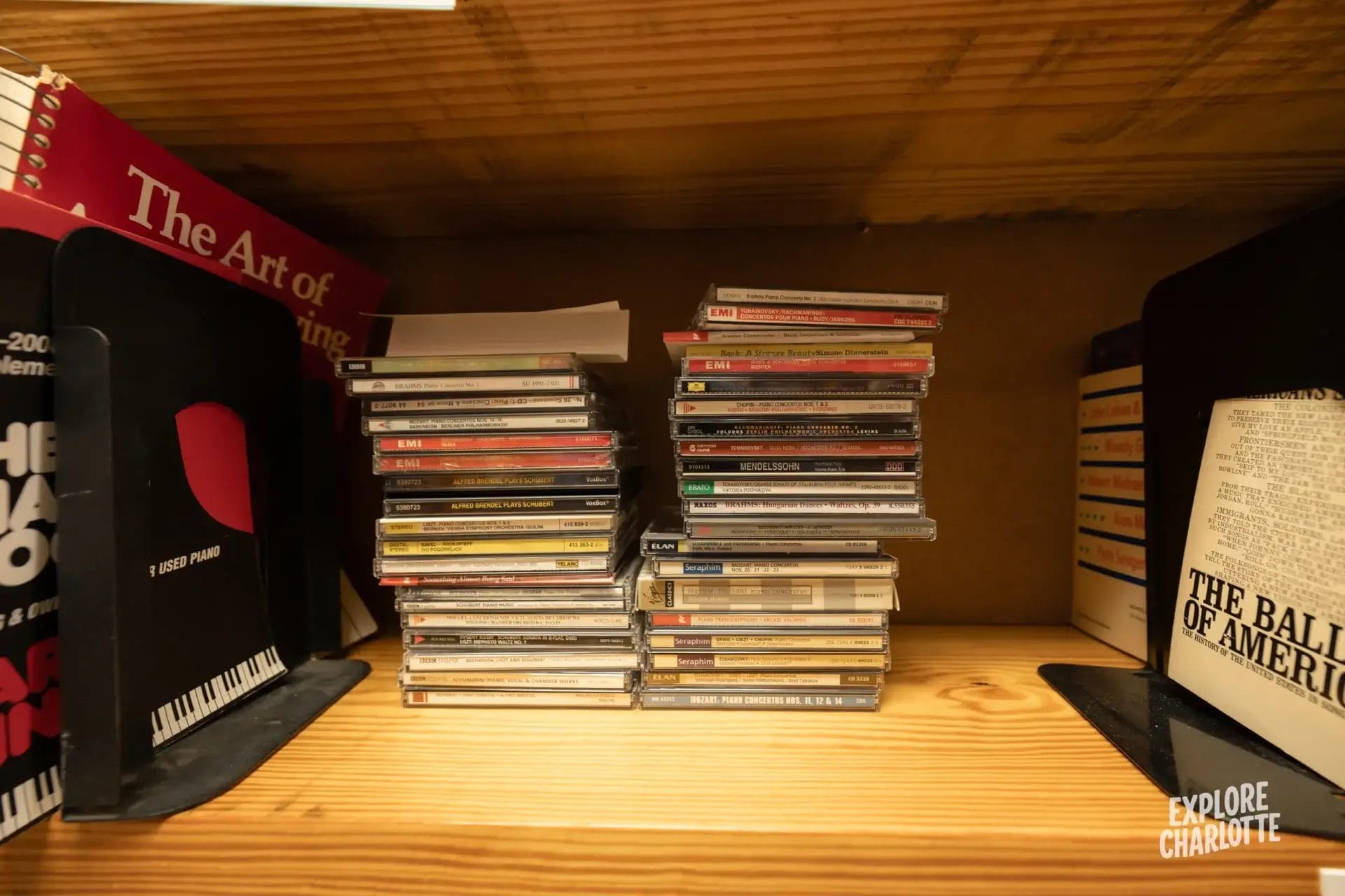 Two stacks of CDs sit on a wooden shelf beside books and a spiral notebook, with "Explore Charlotte" shown in the corner.