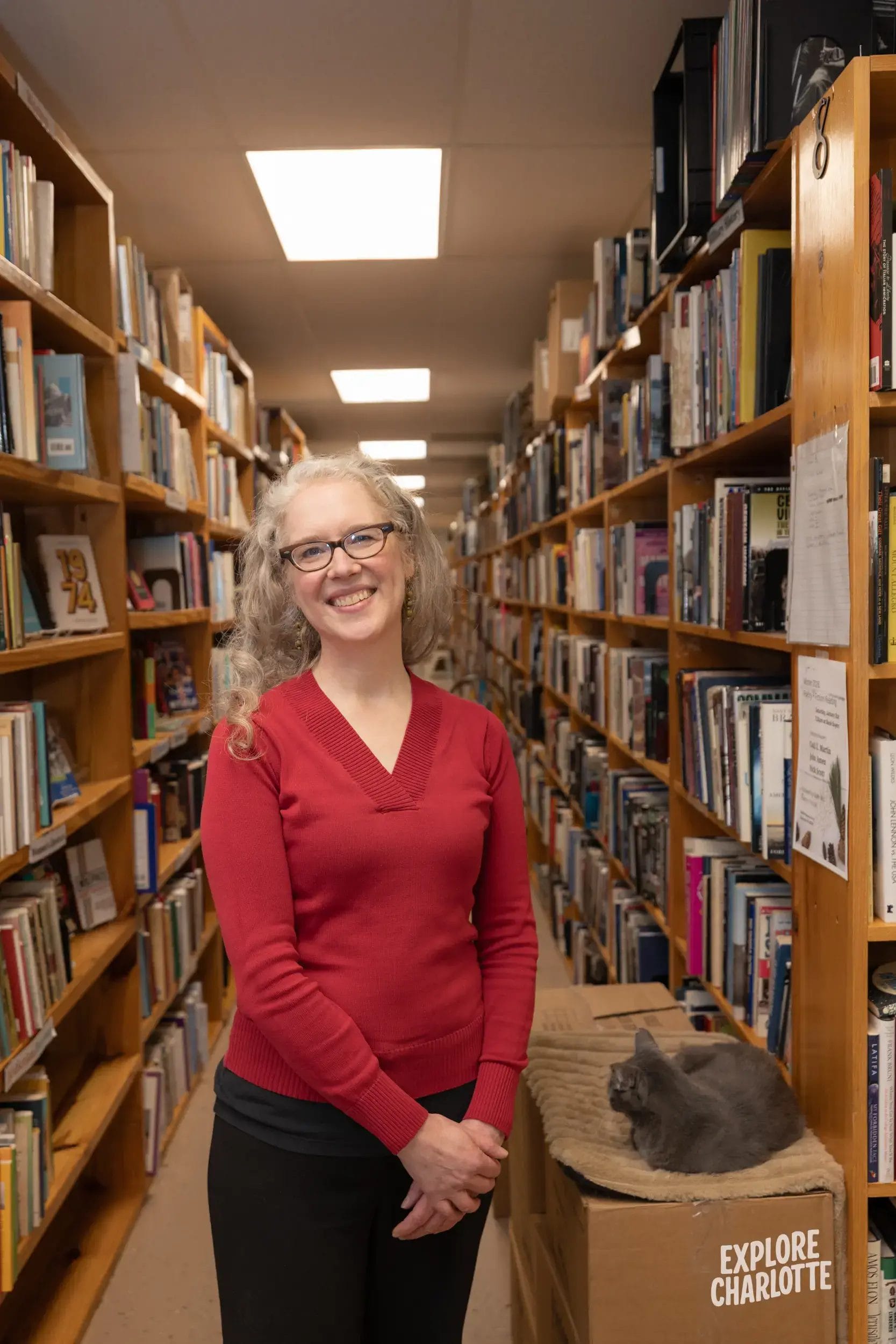 A woman in glasses and a red sweater stands smiling in a Charlotte library aisle. A gray cat lounges on an "Explore Charlotte" box.