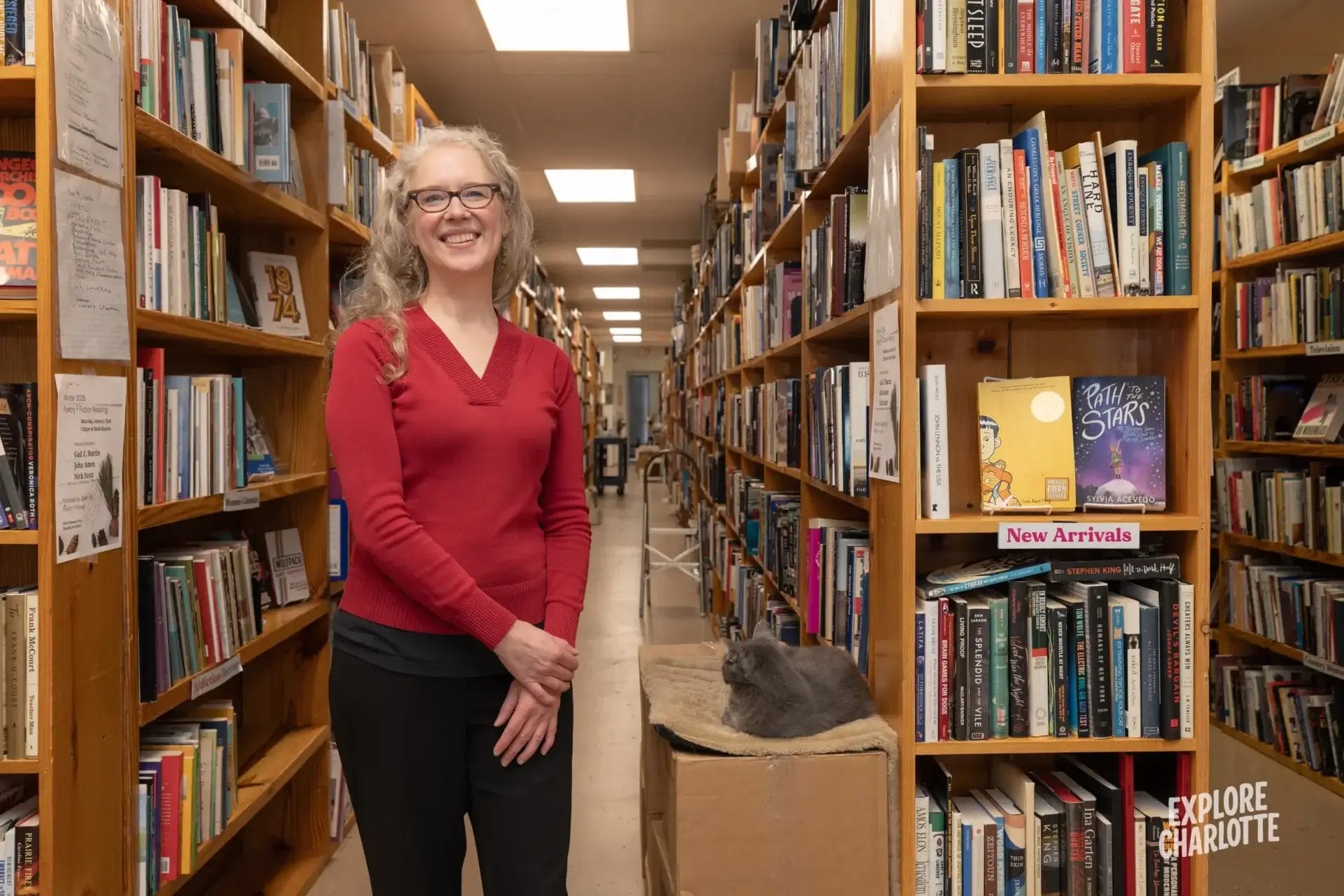 A woman with gray hair and glasses smiles in a Charlotte bookstore aisle, near bookshelves and a gray cat on a cardboard box.