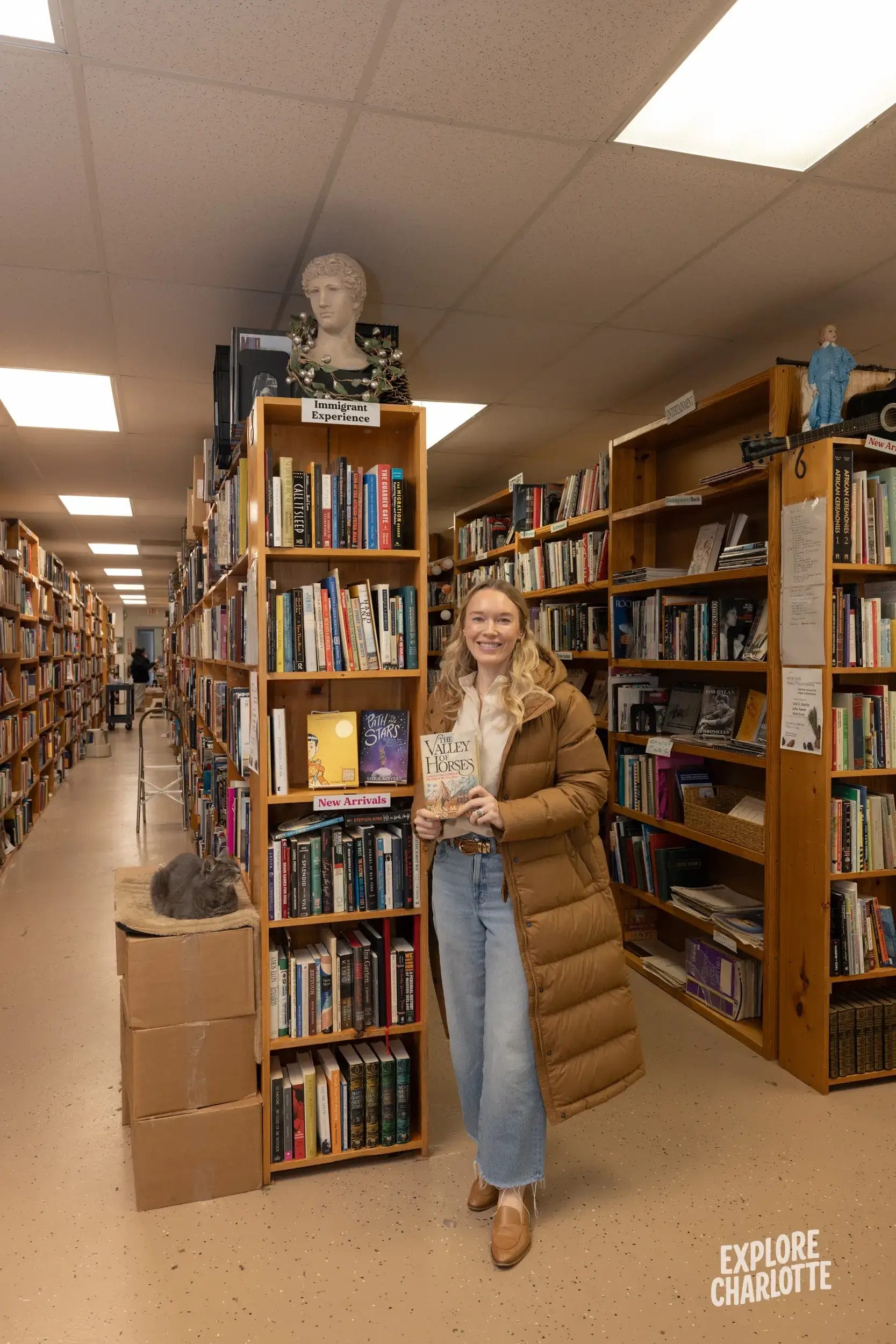 A smiling woman in a brown coat holds a book in a Charlotte bookstore. A cat naps on a box; shelves and a bust fill the cozy scene.