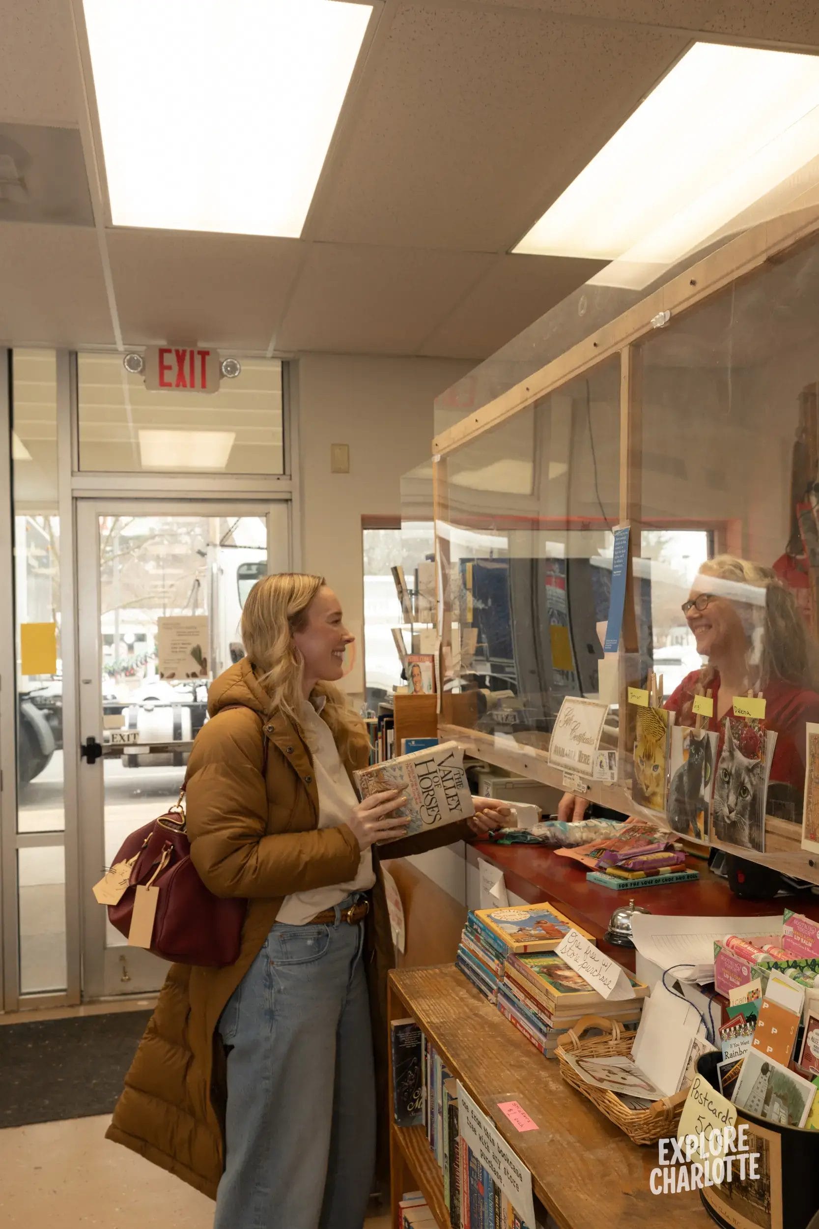 A woman in a brown coat talks with a staff member at a Charlotte library checkout counter. Book shelves line the area beside her.