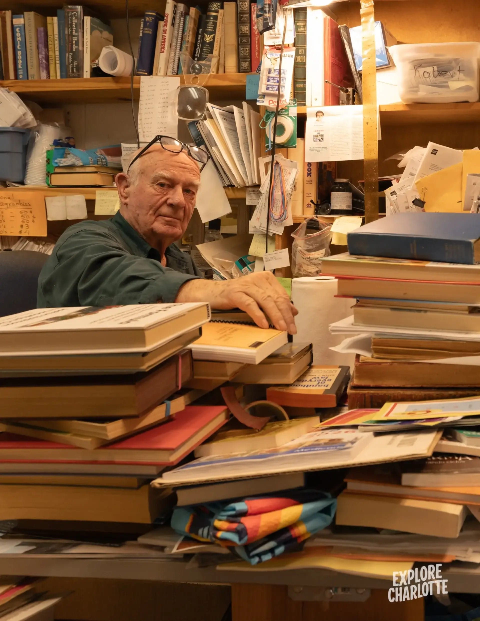 An older man sits at a cluttered desk in Inside Book Buyers, a longtime Charlotte used bookstore, books piled high around him.