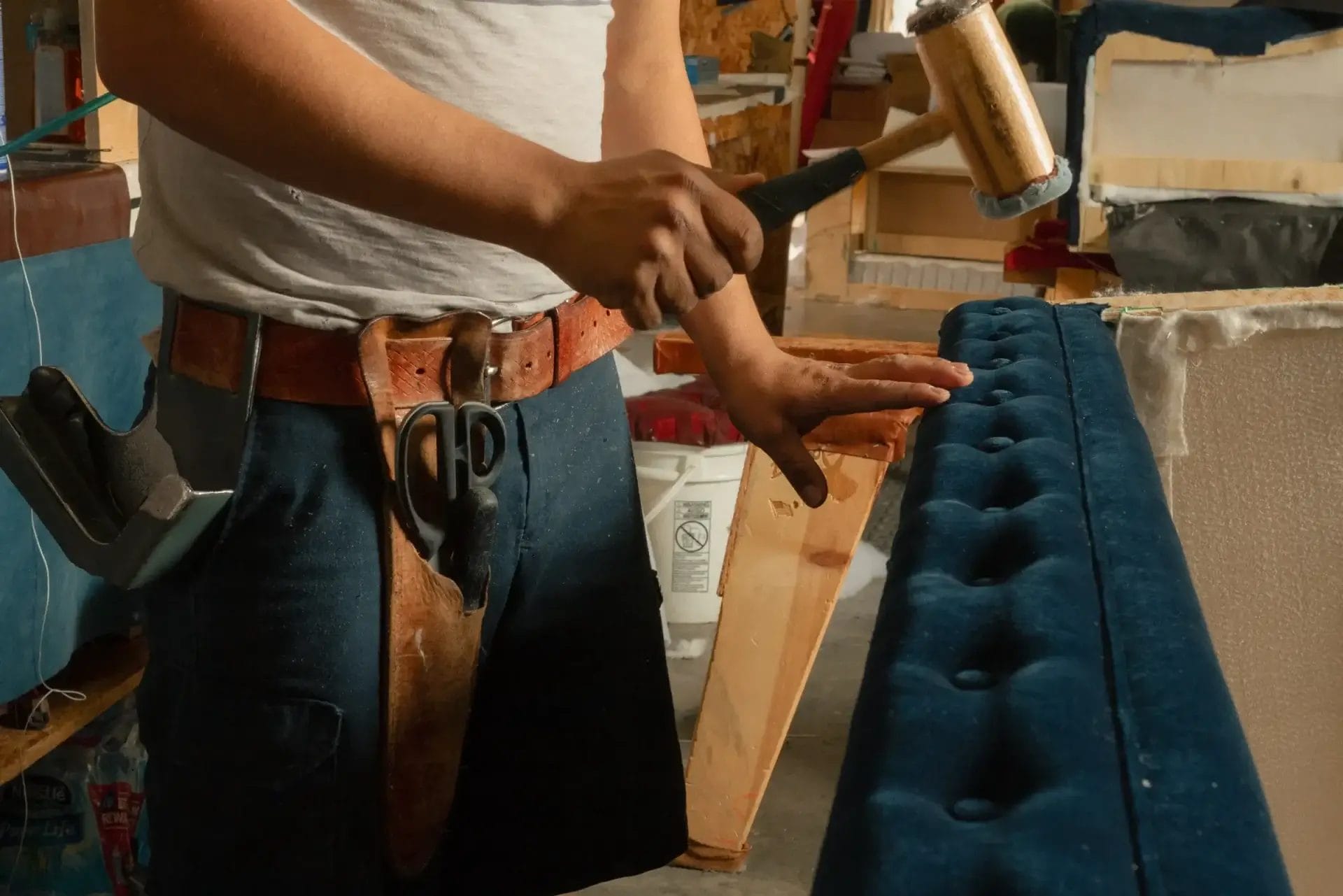Someone in a tool belt works on upholstering a blue, tufted chair at COCOCO Home’s workshop in North Carolina.