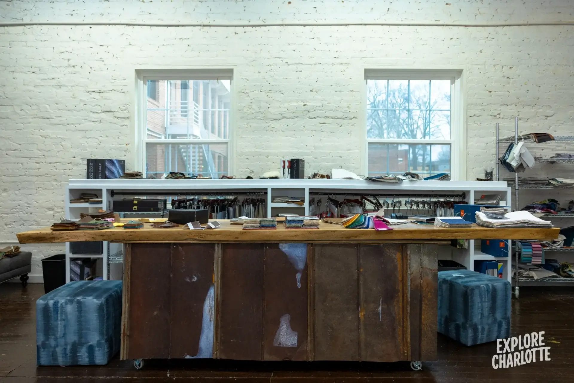 Cozy workspace with a wooden table, fabric swatches, and books by a white brick wall—“Explore Charlotte” shows in the corner.