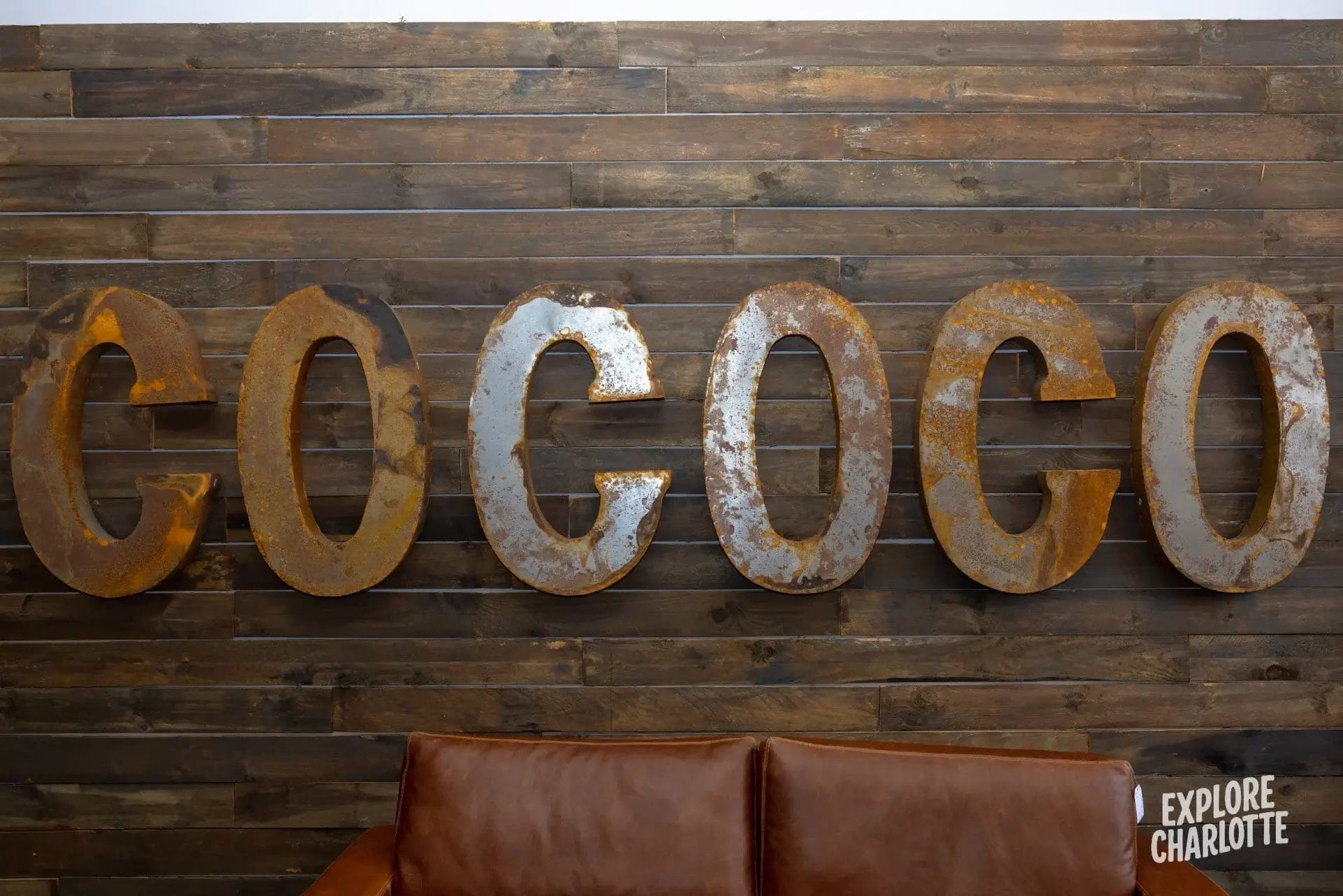 Rusty metal letters spelling "COCOCO" hang above a brown leather couch; "Explore Charlotte" appears in white at the corner.