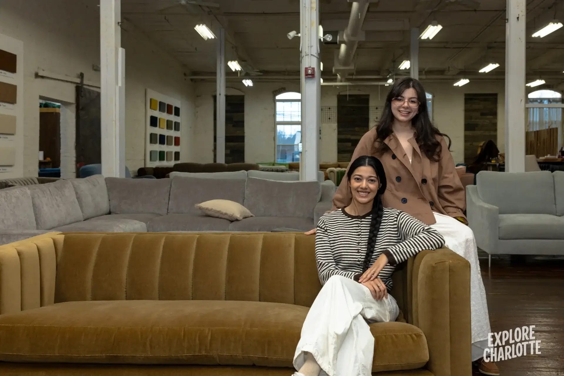 Two women smile together in a Charlotte COCOCOHome showroom, with cozy sofas and fabrics around. "Explore Charlotte" is shown.