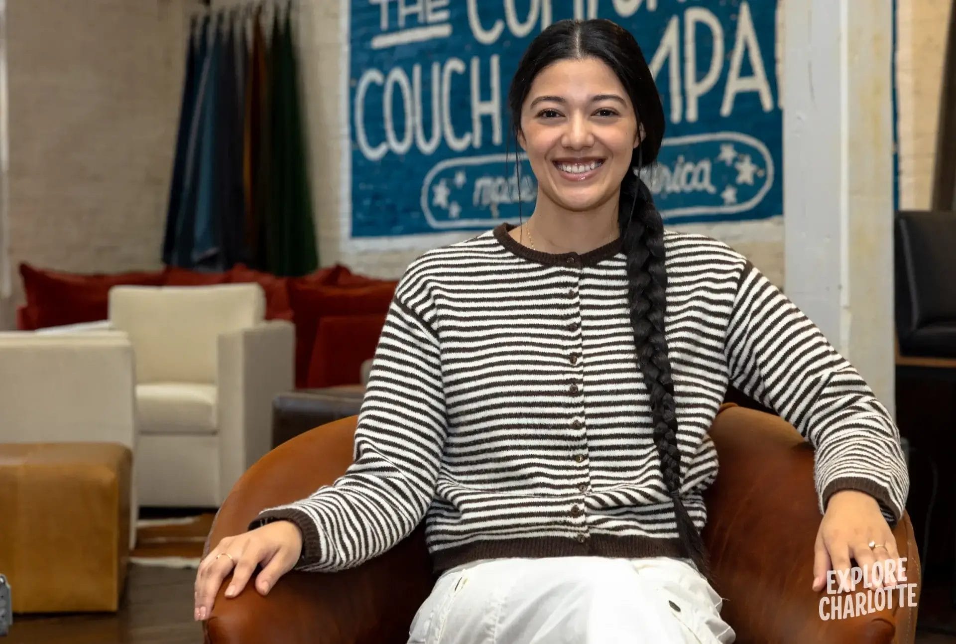 A woman in a striped sweater and white pants smiles, sitting on COCOCOHome furniture with "THE COUCH" and "EXPLORE CHARLOTTE" signs.