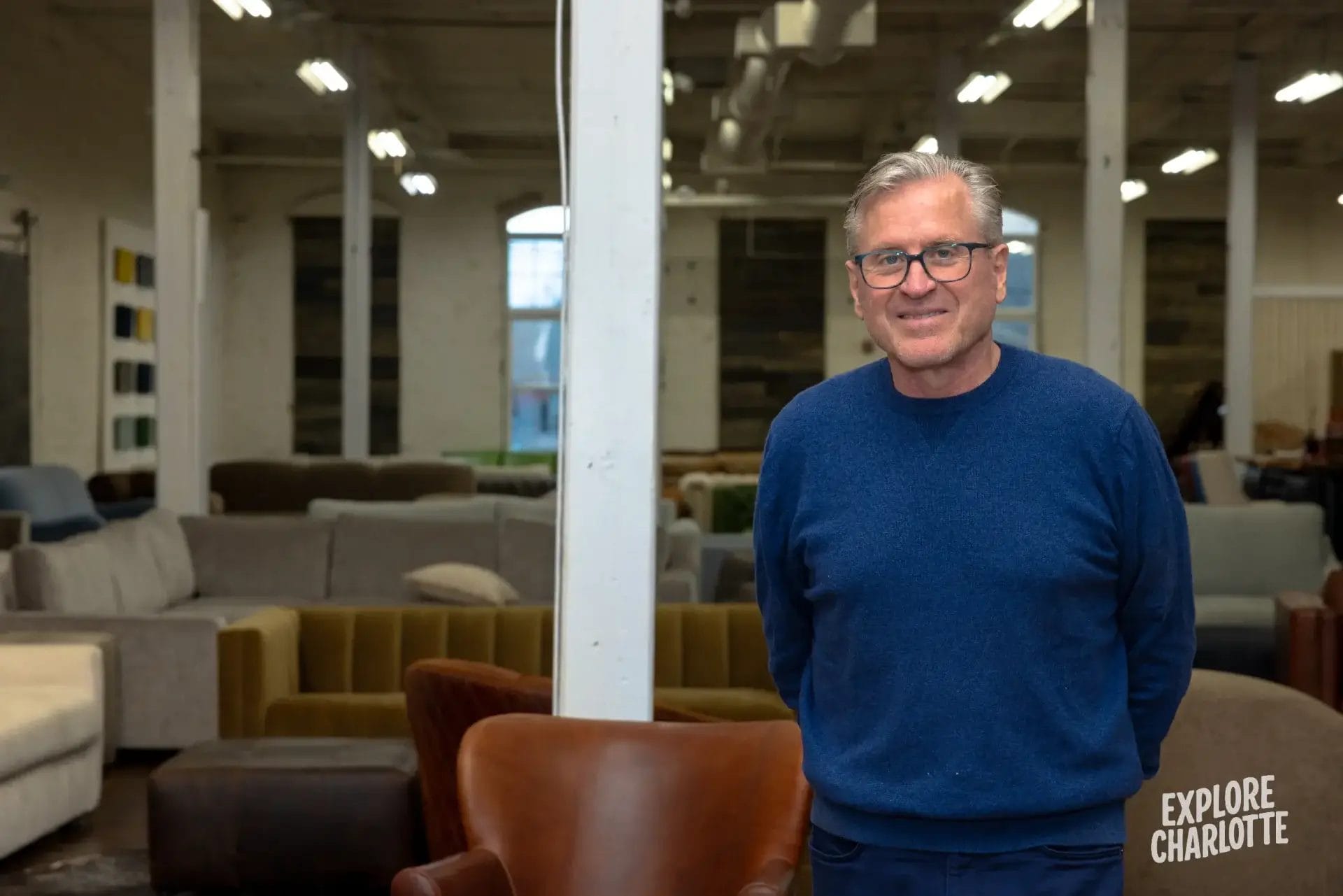 A smiling man in glasses and a blue sweater stands in COCOCOHome’s Charlotte showroom, with sofas and chairs around him. "Explore Charlotte" appears in the corner.