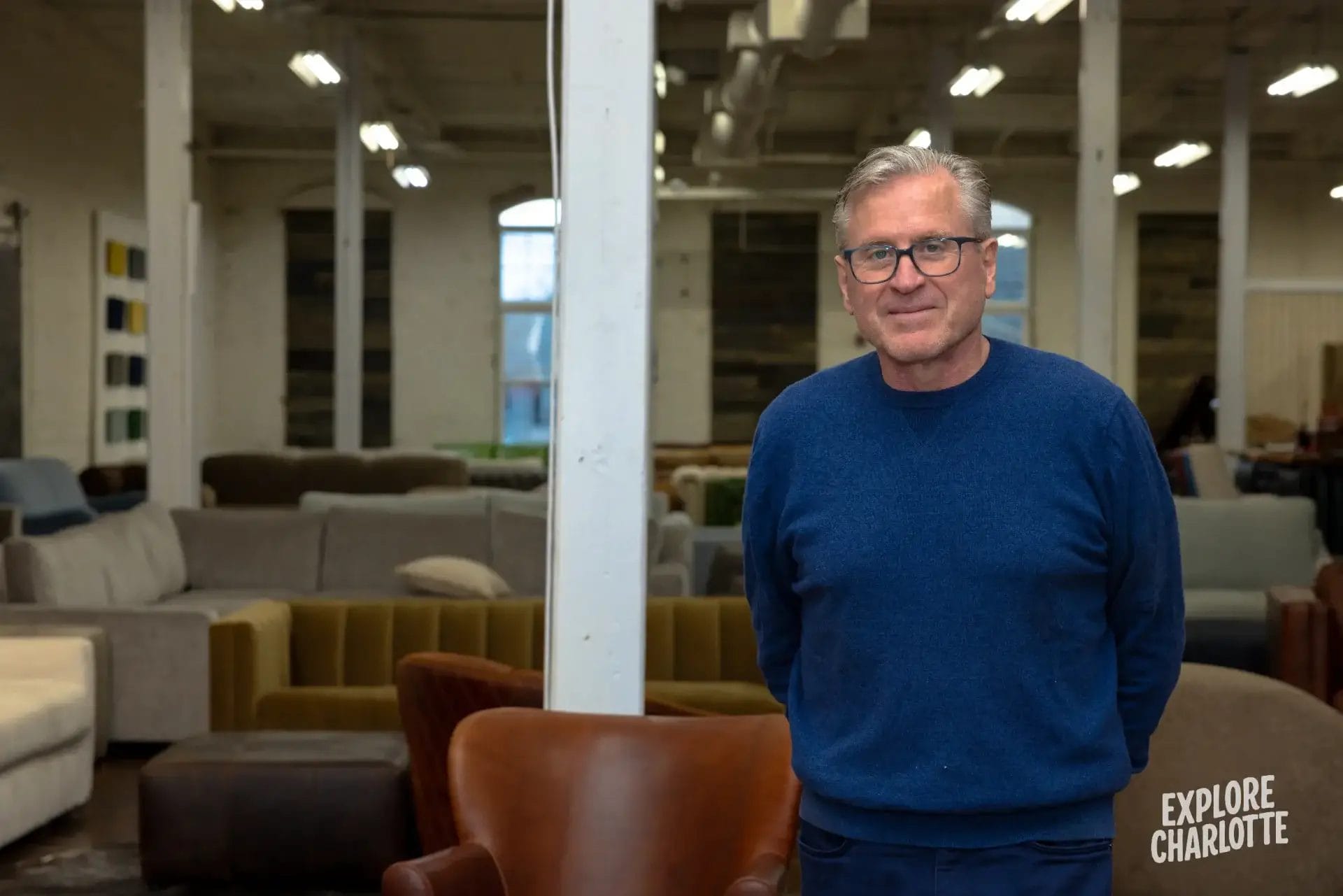 A gray-haired man in glasses and a blue sweater stands in COCOCOHome’s airy showroom of sofas. “Explore Charlotte” is shown.
