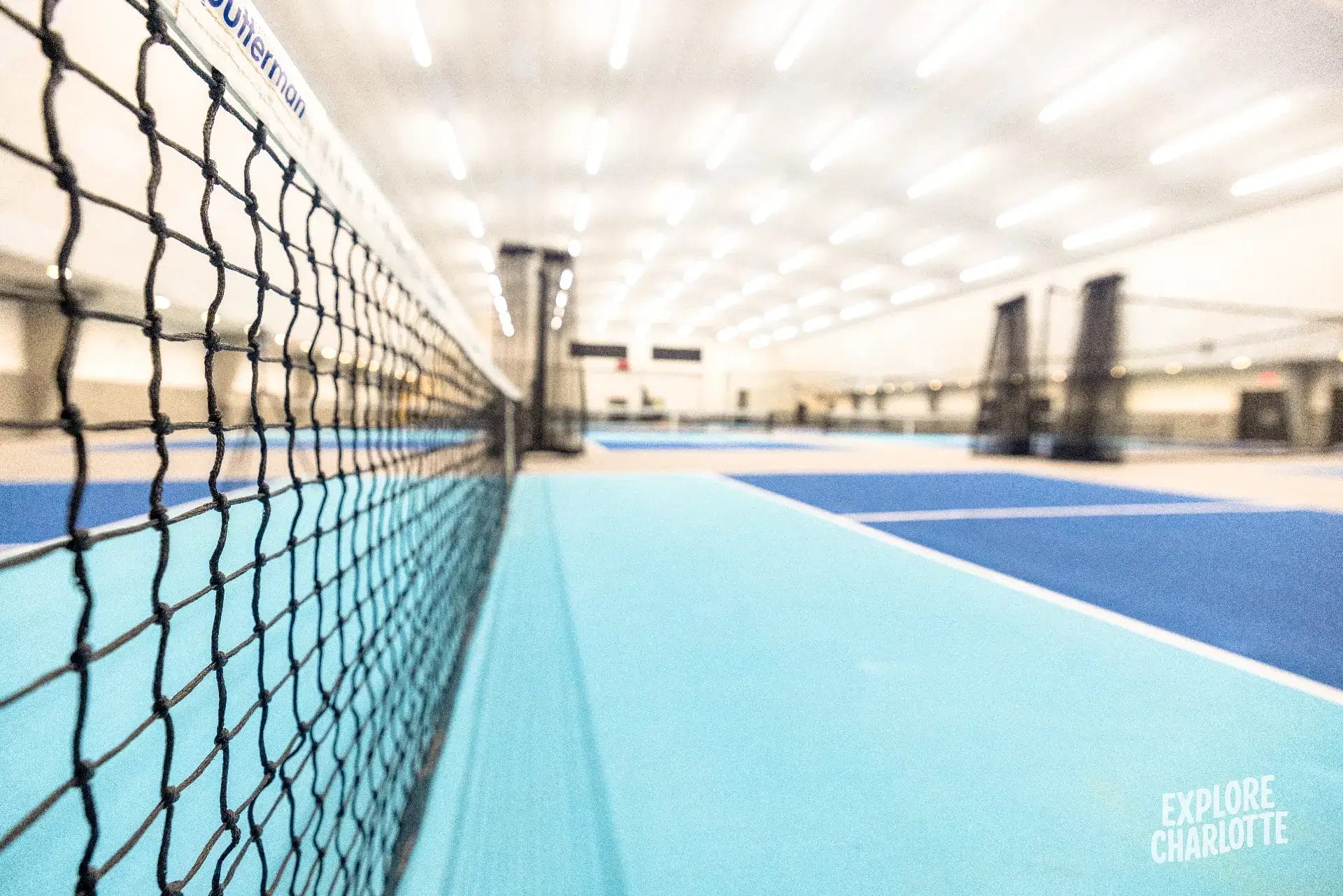 Low-angle shot of an indoor Charlotte tennis court with a black net, blue lines, bright lights, and "Explore Charlotte" in view.