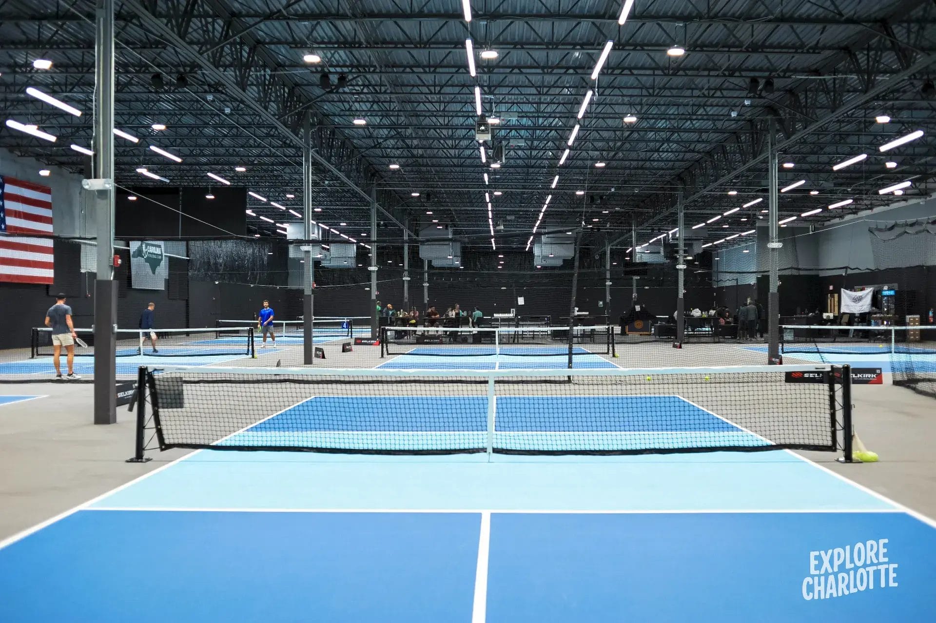 Indoor pickleball courts at Carolina Pickleball Club in Charlotte, with locals playing under bright lights and "EXPLORE CHARLOTTE" on the floor.