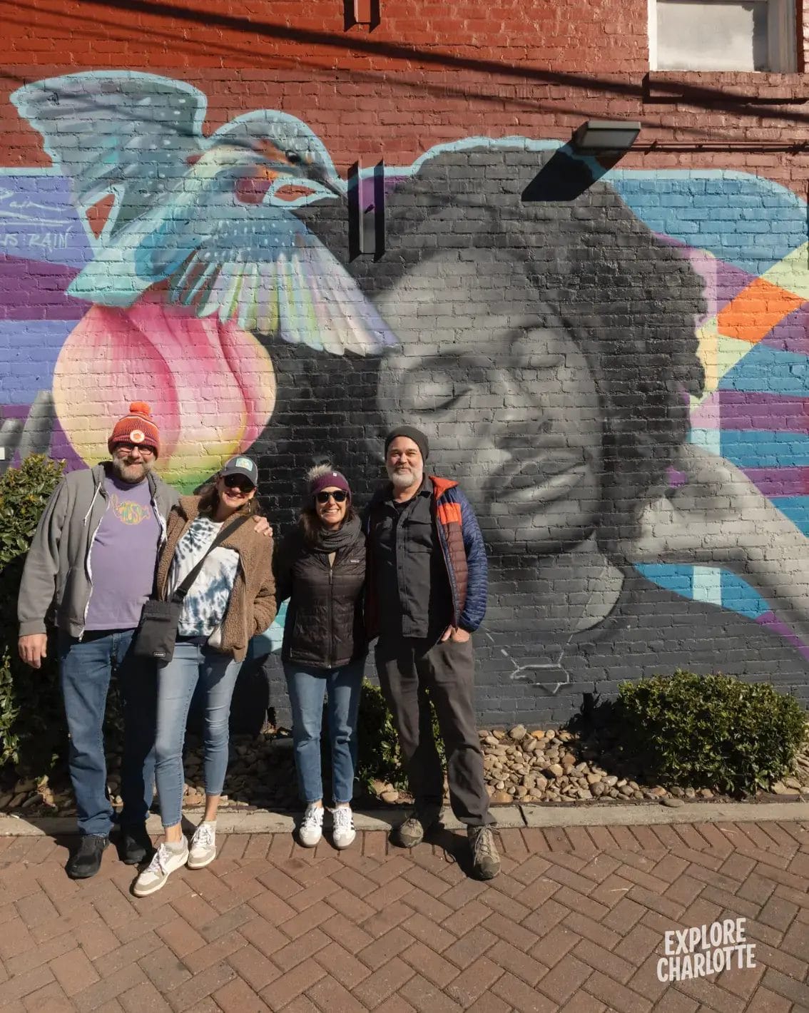 Four people stand by a vibrant Charlotte mural on brick, showing a serene face, bird, peach, and "EXPLORE CHARLOTTE" in the corner.
